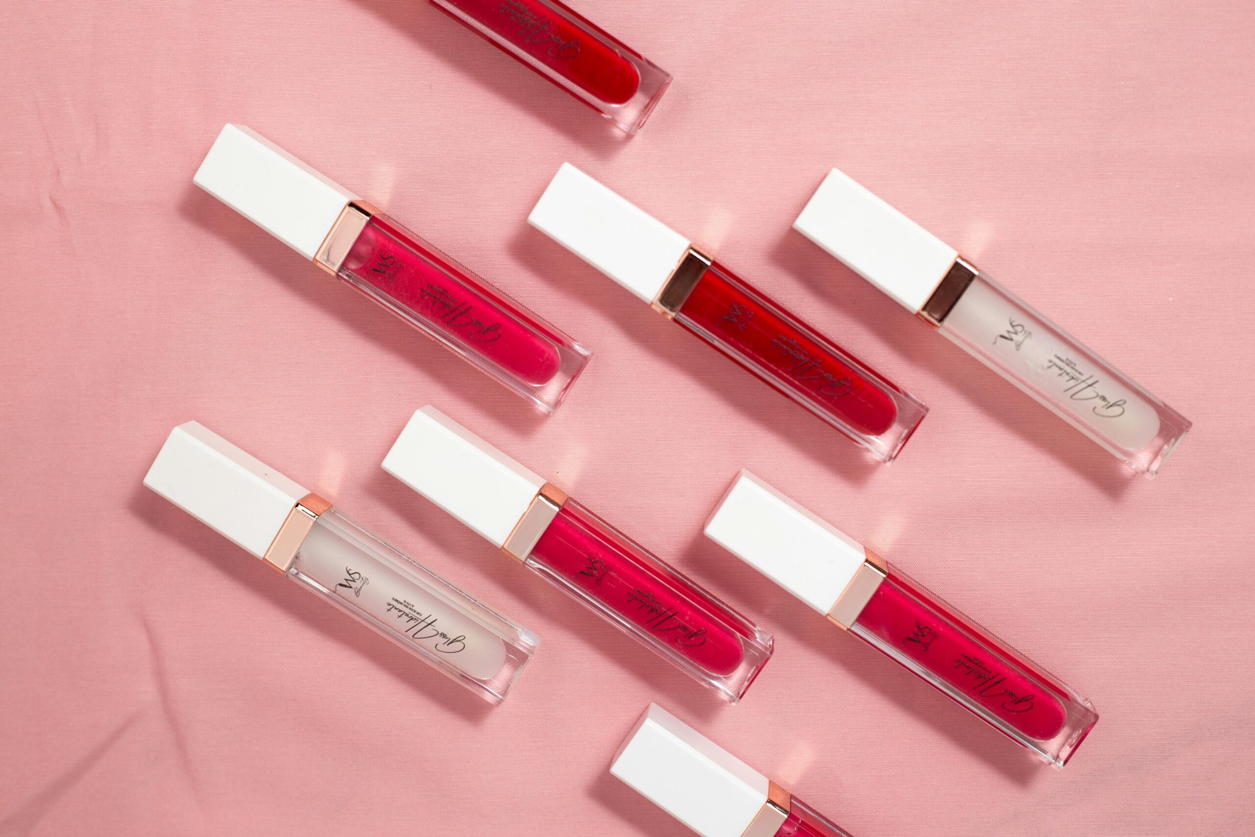 Flat lay of colorful lip glosses on a soft pink fabric background.